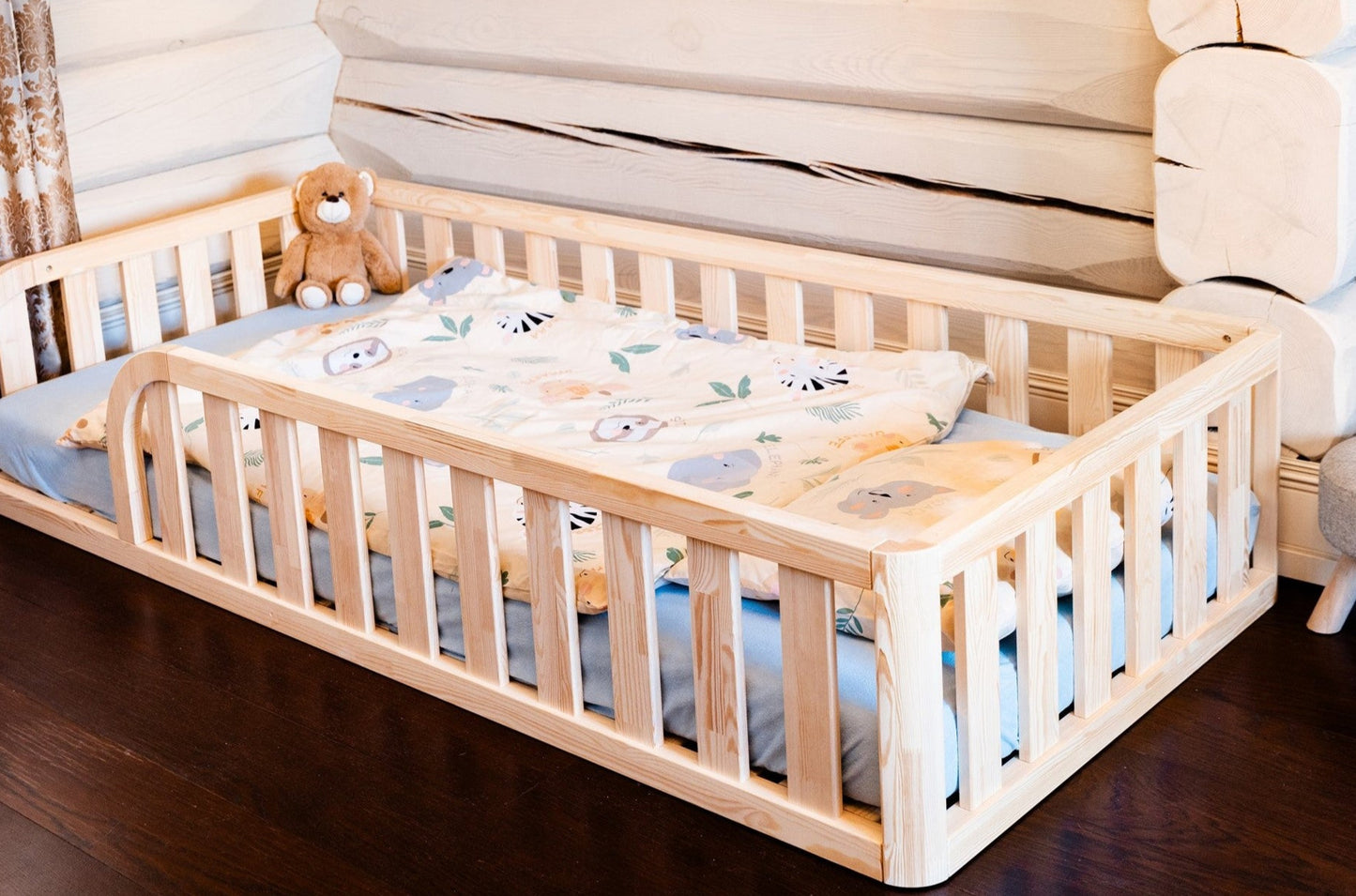 Wooden Montessori toddler floor bed (bed with slats and rails and with rounded opening for kids safety). The bed sits on a floor in a room. And there is mattress in the bed. Bed is with rounded outer corners.