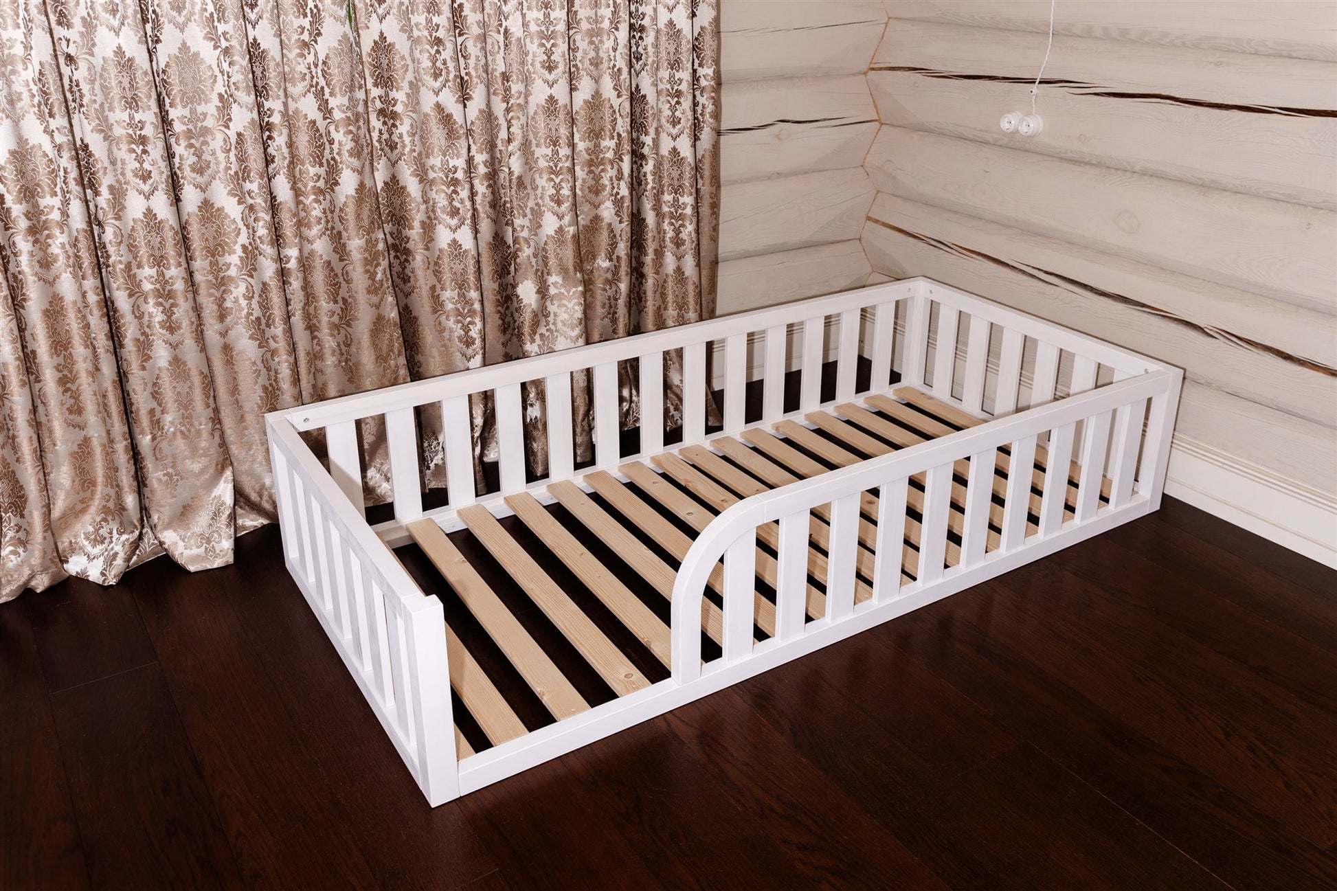 Wooden Montessori toddler floor bed (bed with slats and rails and with rounded opening for kids safety). The bed sits on a floor in a room. White bed frame.