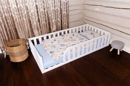 Wooden Montessori toddler floor bed (bed with slats and rails and with rounded opening for kids safety). The bed sits on a floor in a room. And there is mattress in the bed. White bed.