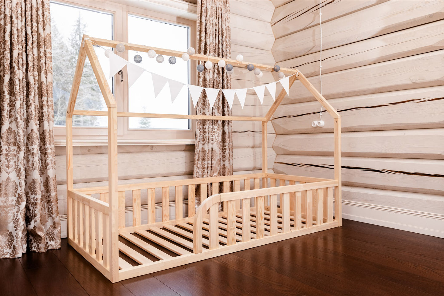 Wooden toddler house floor bed (bed with a house-shaped frame and a cut out roof, with slats and rails and with rounded opening for kids safety). The bed sits on a floor in a room with a window.