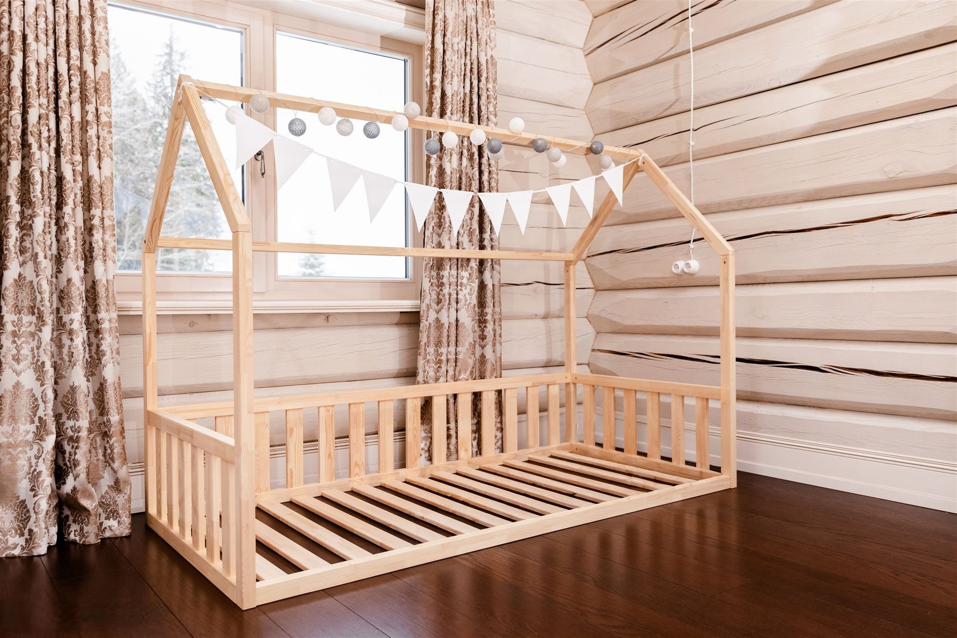 Wooden toddler house floor bed (bed with a house-shaped frame and a cut out roof, with slats and rails, but without front rails). The bed sits on a floor in a room with a window.