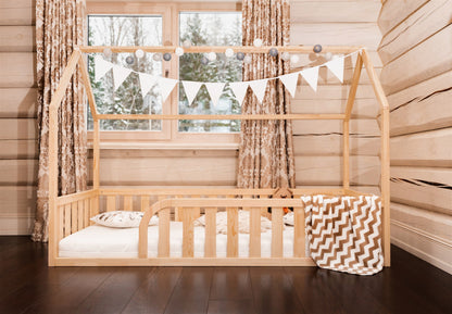 Wooden toddler house floor bed (bed with a house-shaped frame and a cut out roof, with slats and rails and with rounded opening for kids safety). The bed sits on a floor in a room with a window. And there is mattress and teddy bear in the bed.