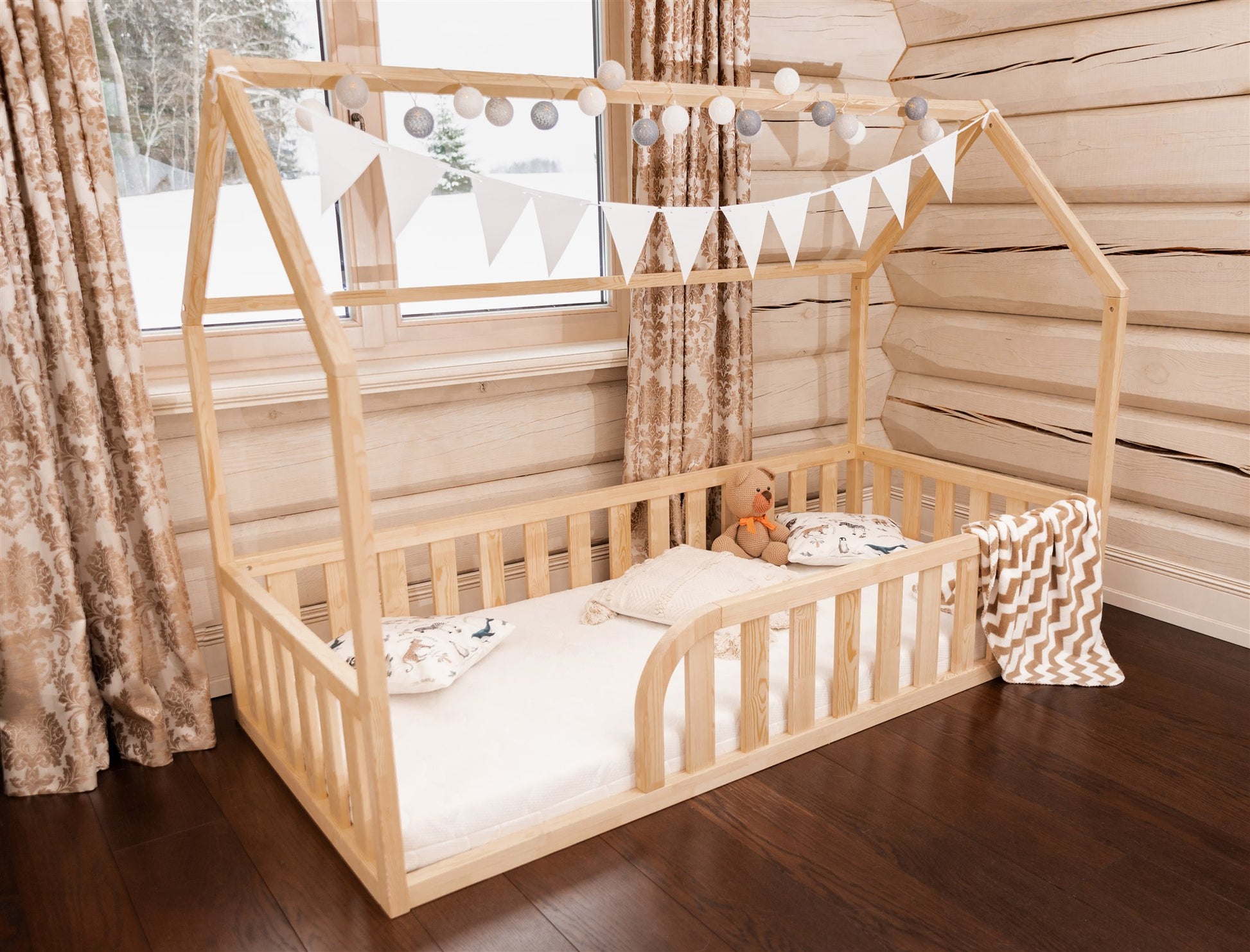 Wooden toddler house floor bed (bed with a house-shaped frame and a cut out roof, with slats and rails and with rounded opening for kids safety). The bed sits on a floor in a room with a window. And there is mattress and teddy bear in the bed.