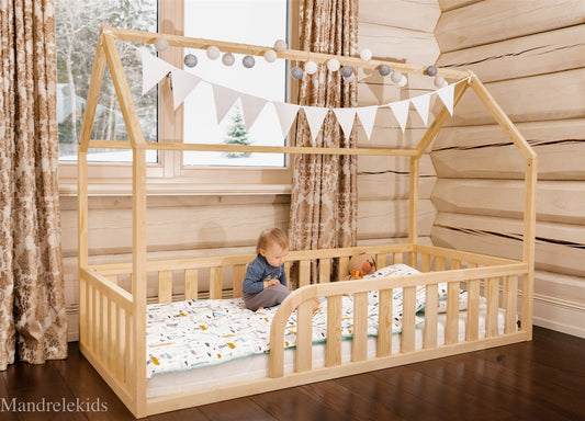 Wooden toddler house floor bed (bed with a house-shaped frame and a cut out roof, with slats and rails and with rounded opening for kids safety). The bed sits on a floor in a room with a window. And there is toddler and teddy bear sitting on the mattress in the bed.