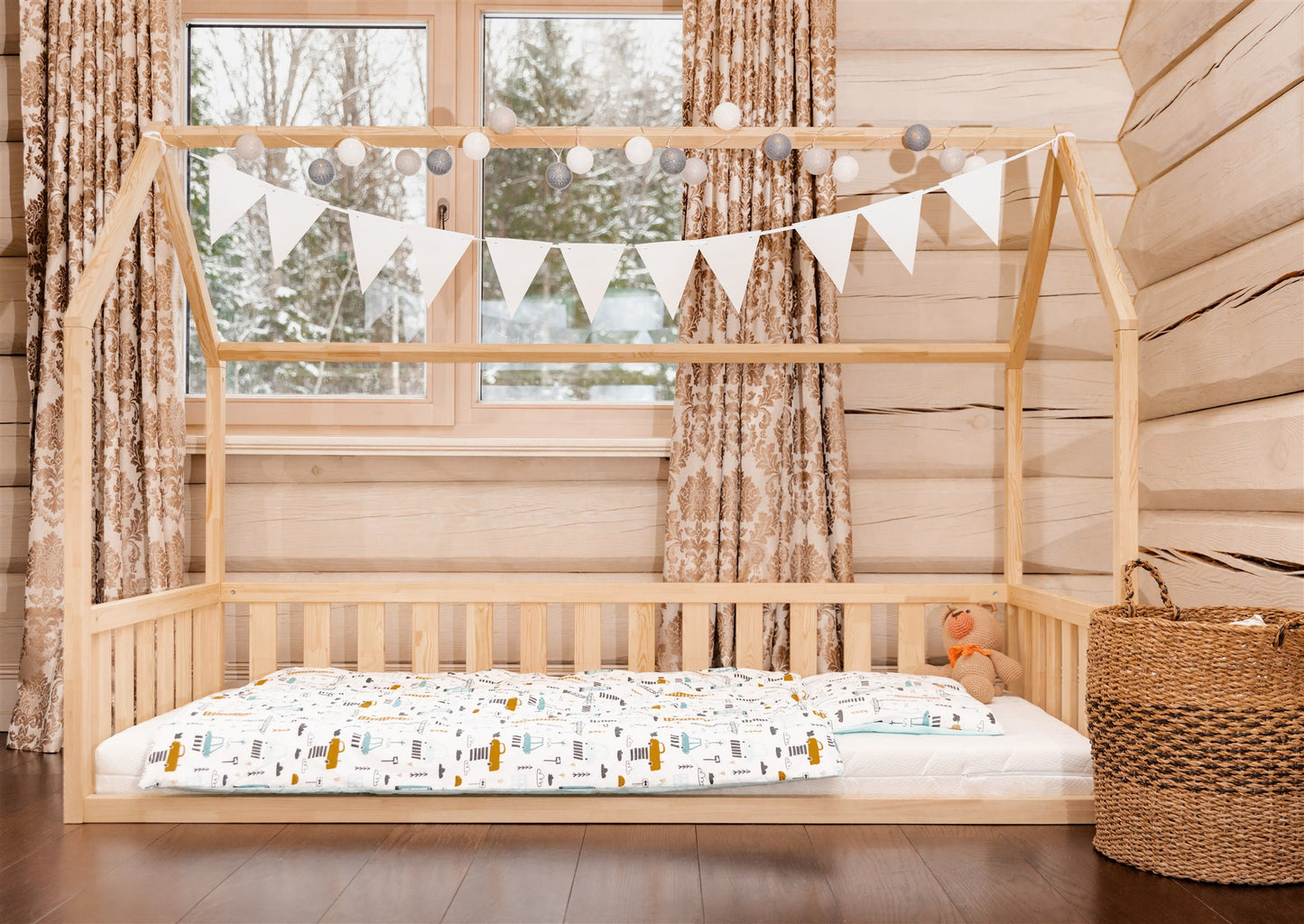 Wooden toddler house floor bed (bed with a house-shaped frame and a cut out roof, with slats and rails, but without front rails). The bed sits on a floor in a room with a window. And there is teddy bear sitting on the mattress in the bed.