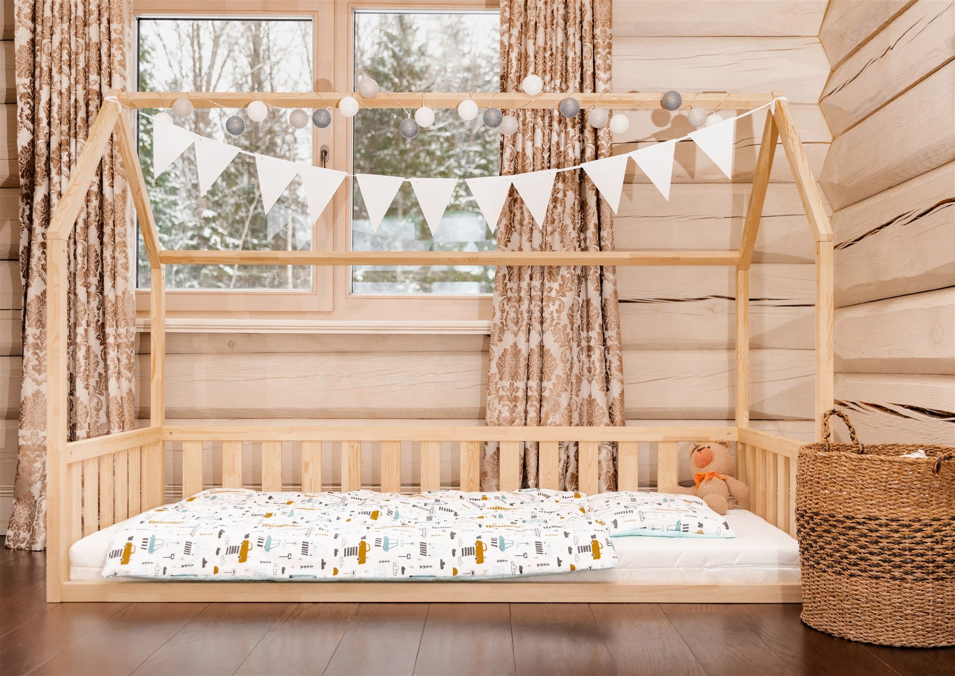 Wooden toddler house floor bed (bed with a house-shaped frame and a cut out roof, with slats and rails, but without front rails). The bed sits on a floor in a room with a window. And there is teddy bear sitting on the mattress in the bed.