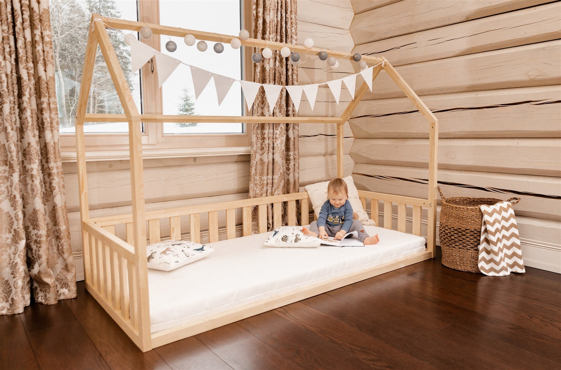 Wooden toddler house floor bed (bed with a house-shaped frame and a cut out roof, with slats and rails, but without front rails). The bed sits on a floor in a room with a window. And there is toddler sitting on the mattress in the bed and reading a book.