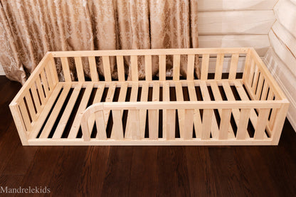 Wooden Montessori toddler floor bed (bed with slats and rails and with rounded opening for kids safety). The bed sits on a floor in a room. Natural bed frame.