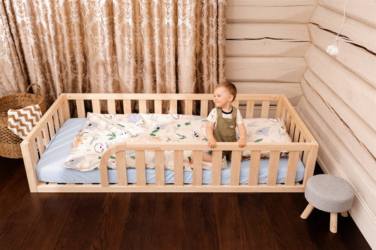 Wooden Montessori toddler floor bed (bed with slats and rails and with rounded opening for kids safety). The bed sits on a floor in a room. And there is toddler sitting on the mattress in the bed.