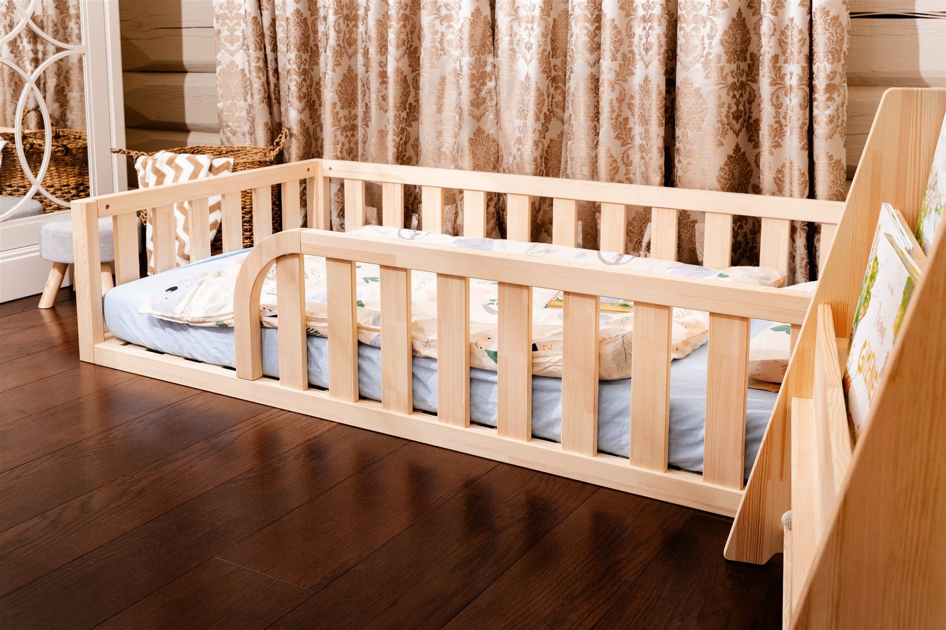 Wooden Montessori toddler floor bed (bed with slats and rails and with rounded opening for kids safety). The bed sits on a floor in a room. And there is mattress in the bed.