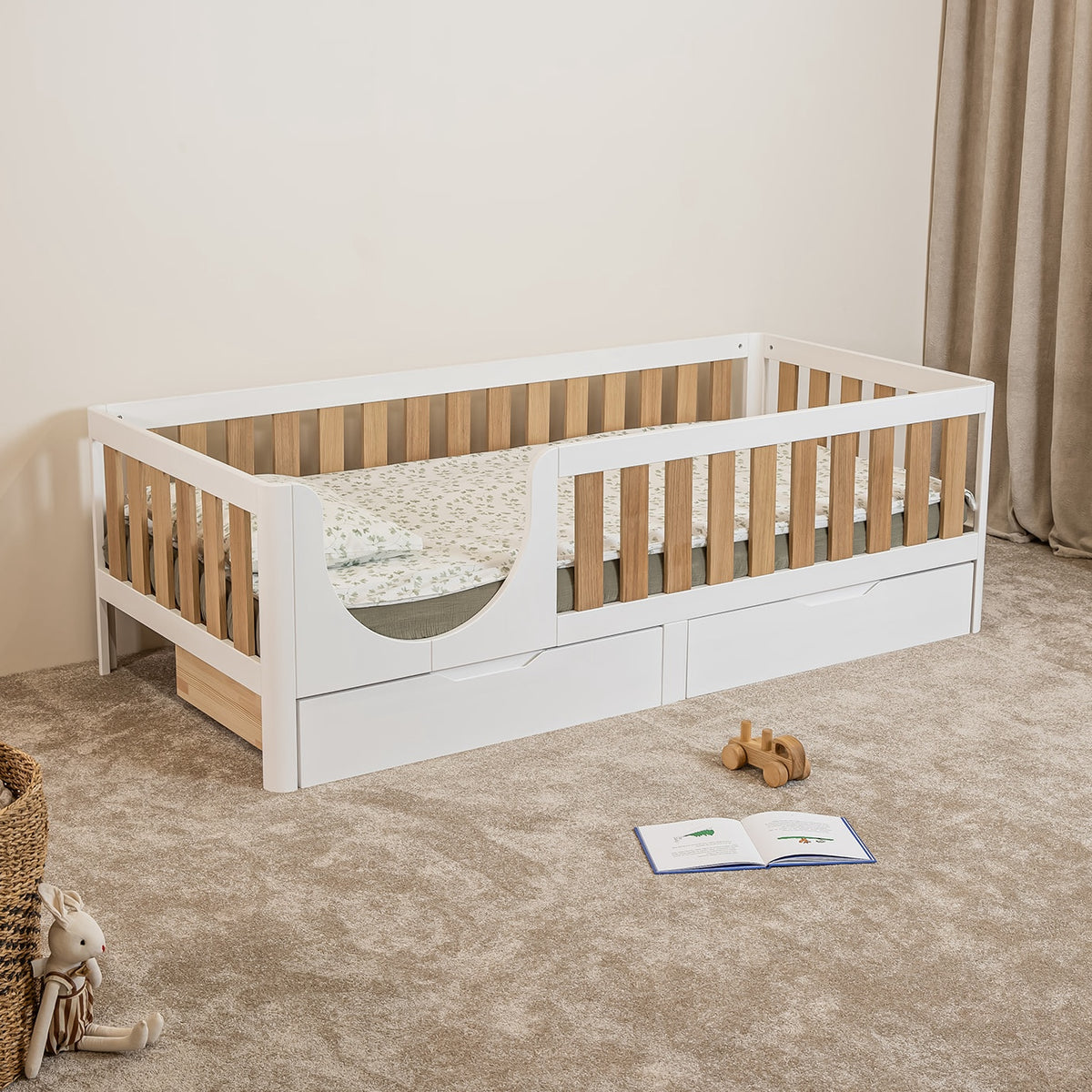 Angled view of EZRA Montessori toddler bed with white frame, oak veneer rails and underbed storage drawers