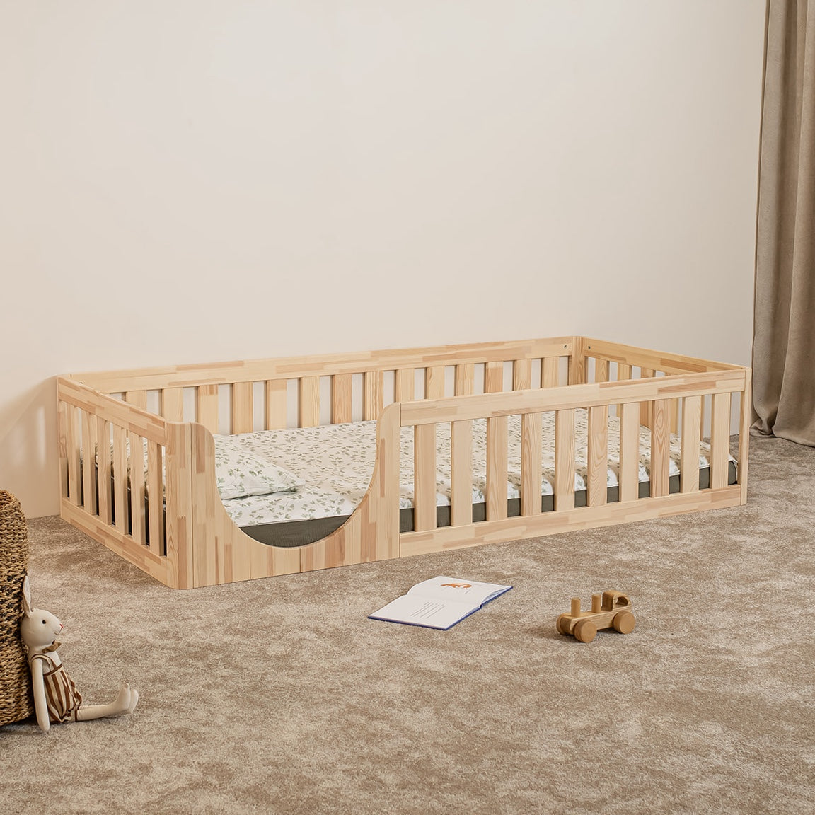 Angled view of EZRA Montessori toddler floor bed with low rounded entrance and natural pine safety rails