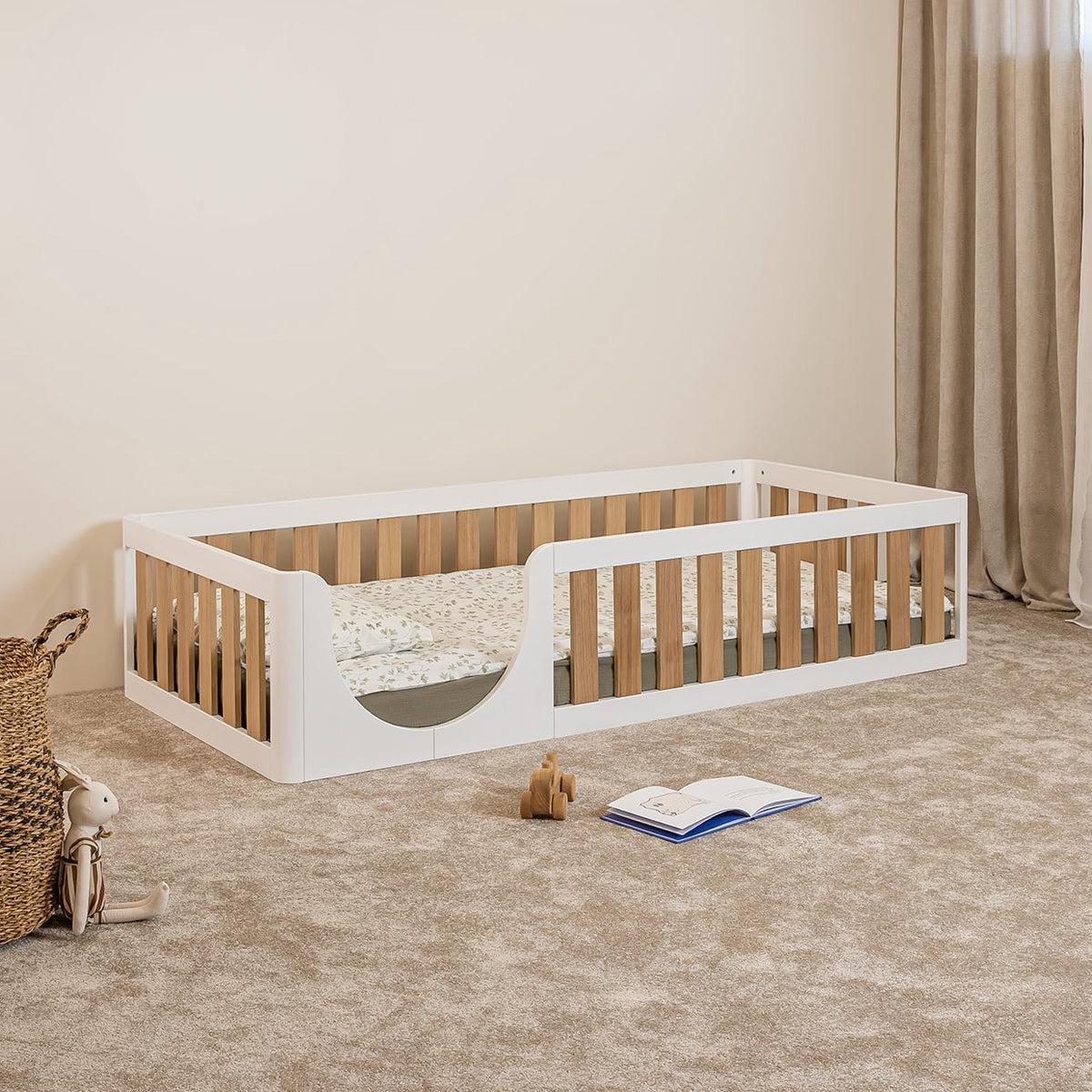 Angled view of EZRA Montessori toddler floor bed with white pine frame, oak veneer safety rails and rounded entrance