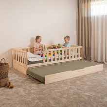 A light natural wood toddler bed with safety rails, shown with an attached olive green storage drawer underneath. Two children are playing on the bed in a softly lit nursery with beige walls, curtains, and carpet.
