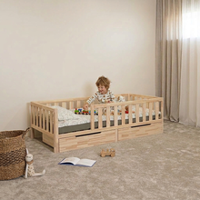 A young child sits in a light natural wood toddler bed with safety rails and under-bed storage drawers, set in a softly lit nursery with beige walls, curtains, carpet, a woven basket, and toys.