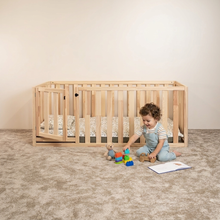 A toddler in light blue overalls sits on a beige carpet next to a natural wood crib, playing with toys. The crib has a patterned mattress and is set against a plain beige wall.