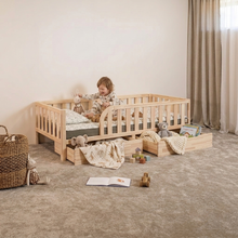 A child sits in a light natural wood toddler bed with safety rails in a softly styled nursery. The bed has built-in storage drawers underneath, holding blankets and toys, with a wicker basket and plush toys on a beige carpet.