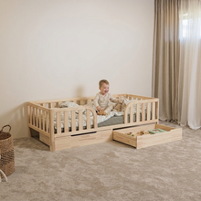 Child sitting on Montessori toddler bed with rounded middle opening and two under-bed storage drawers, one open with toys
