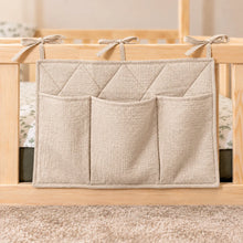 Close-up of a beige quilted cotton bed pocket organiser with three storage pockets and fabric ties on a wooden bed railing.