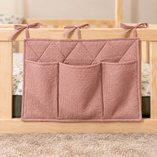 Close-up of a clay pink quilted cotton bed pocket organiser with three storage pockets and fabric ties on a wooden bed railing.