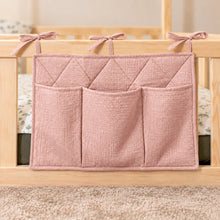 Close-up of a nude pink quilted cotton bed pocket organiser with three storage pockets and fabric ties on a wooden bed railing.