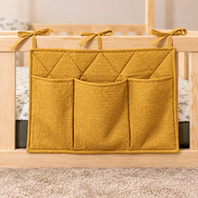 Close-up of a ochre quilted cotton bed pocket organiser with three storage pockets and fabric ties on a wooden bed railing.