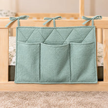 Close-up of a sea green quilted cotton bed pocket organiser with three storage pockets and fabric ties on a wooden bed railing.