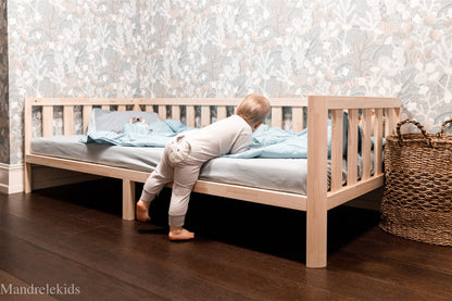 Wooden Montessori toddler bed on legs with rails and a child climbing into bed