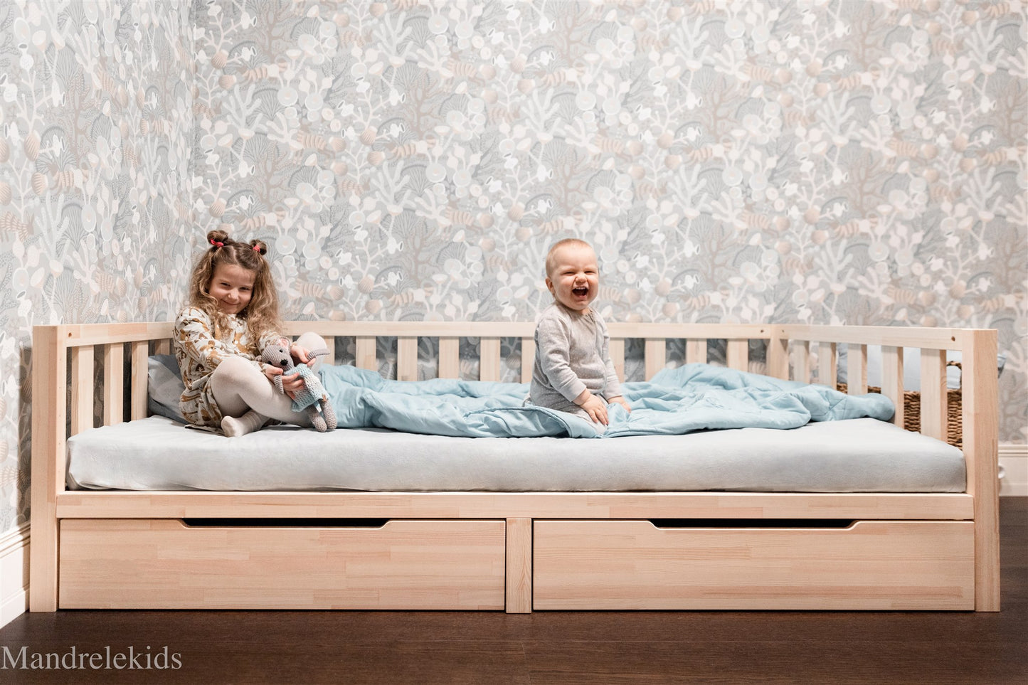 Wooden Montessori toddler bed with storage drawers and two children sitting on the mattress