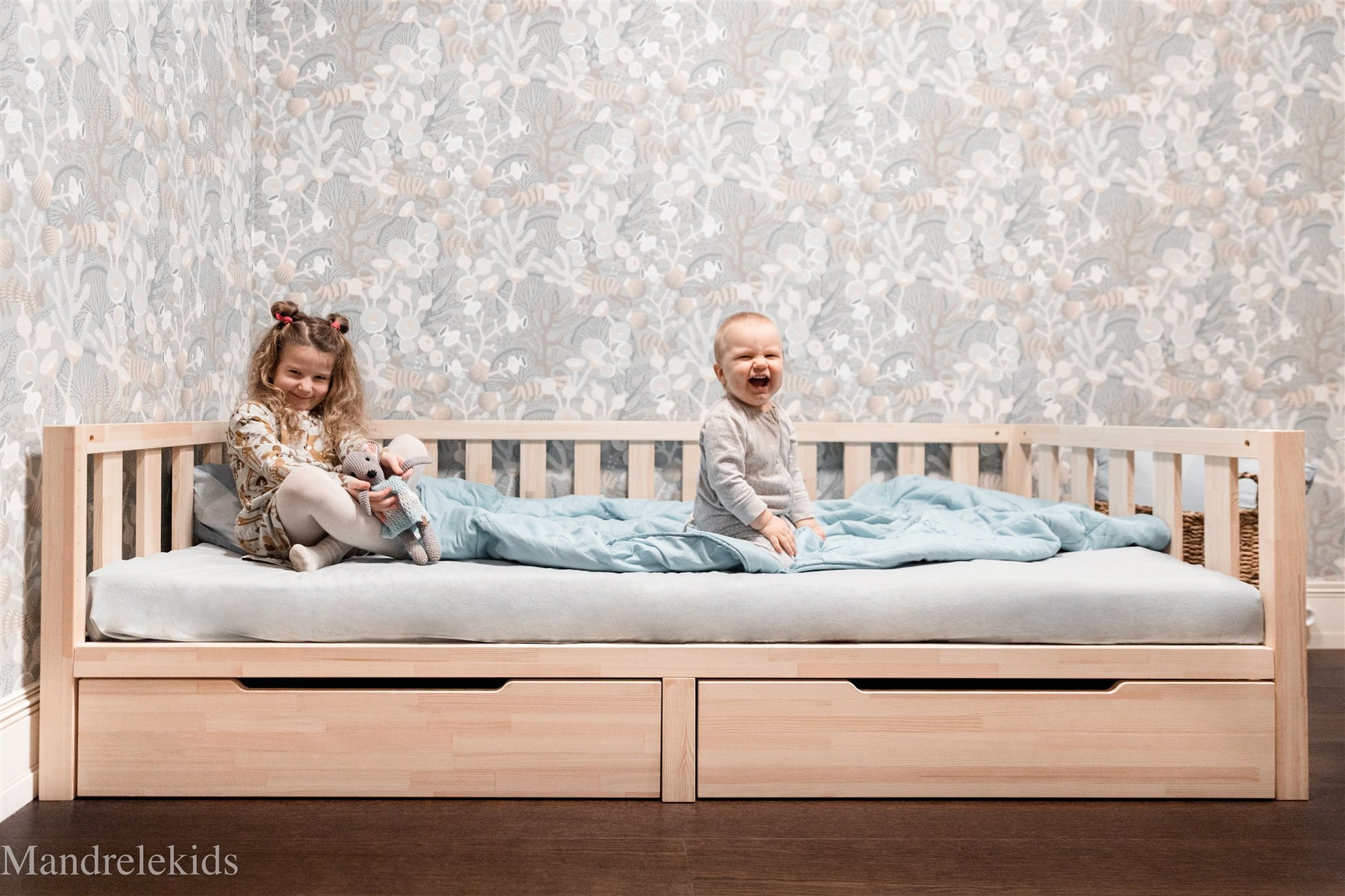 Wooden Montessori toddler bed with storage drawers and two children sitting on the mattress